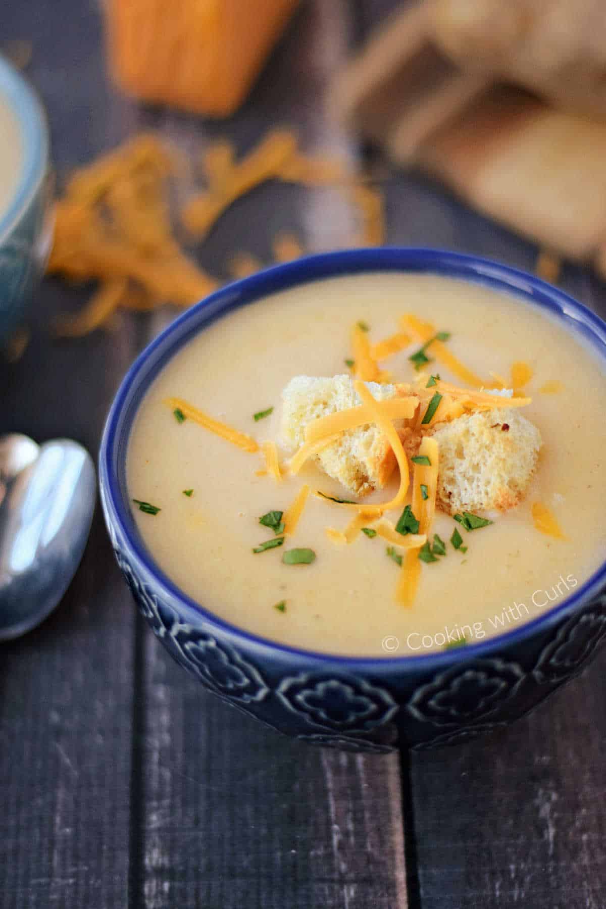 A dark blue bowl filled with potato cheese soup, and topped with croutons and shredded cheddar cheese with additional cheese in the background.