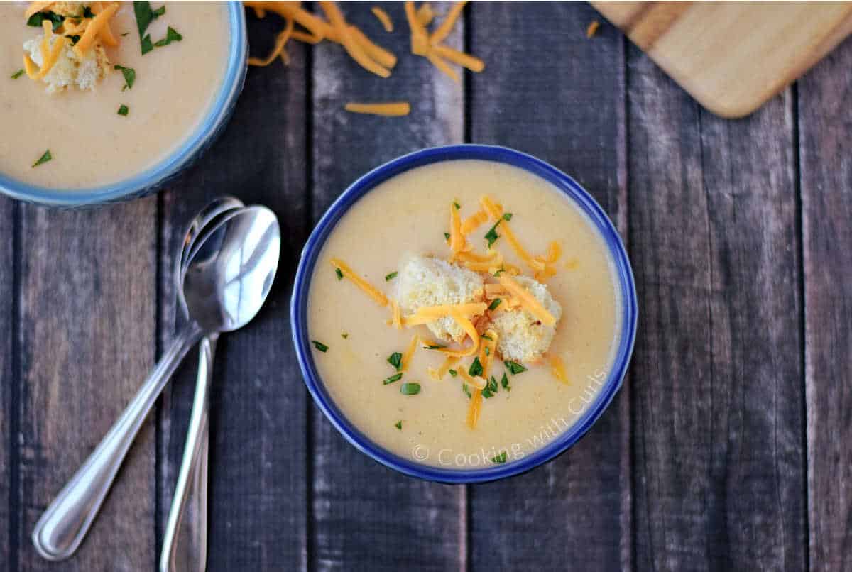 Two blue bowls filled with potato cheese soup sitting on a wooden board with two spoons.