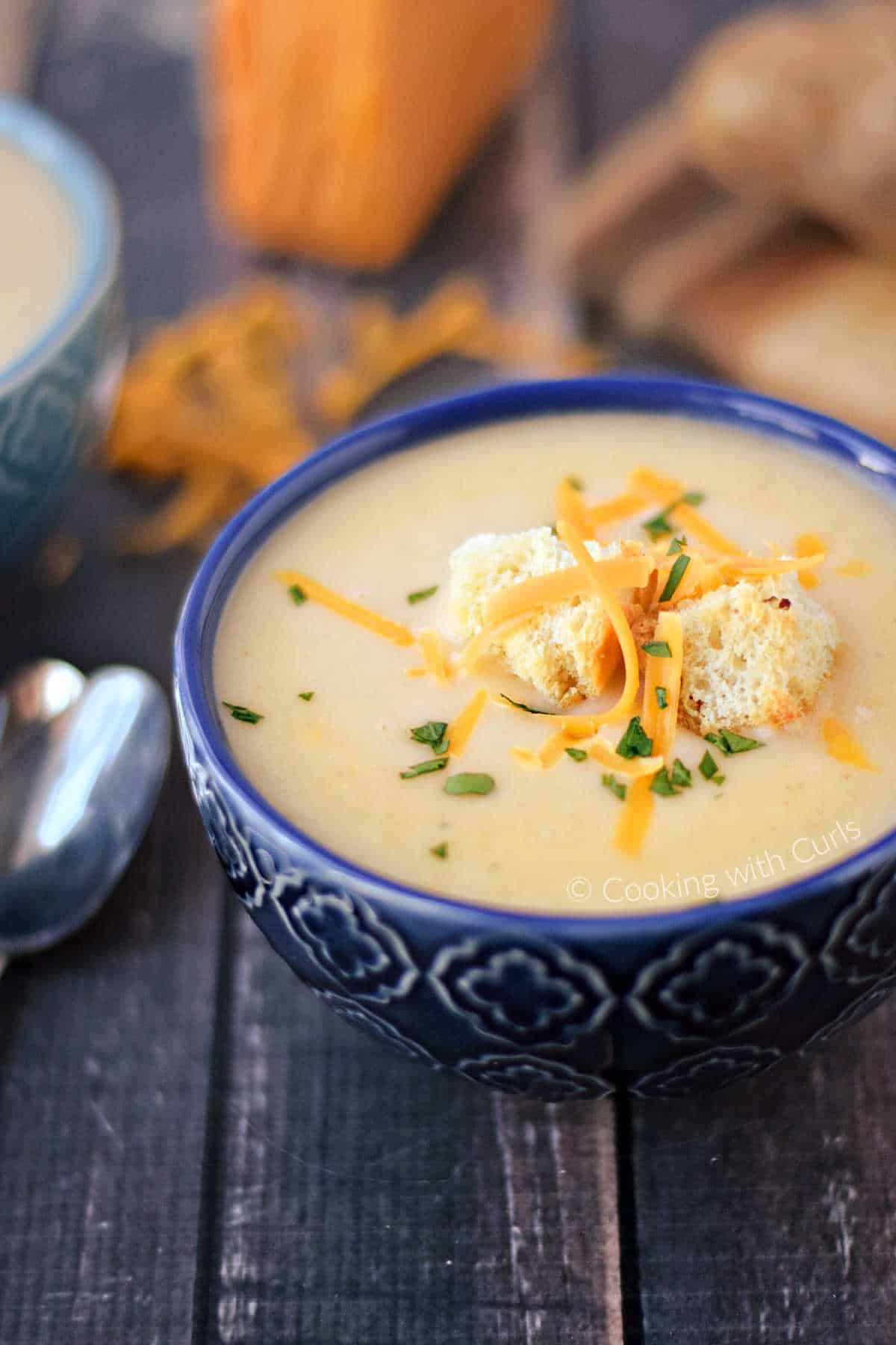 A blue bowl of potato cheese soup and topped with croutons, grated cheddar cheese, and chopped parsley.