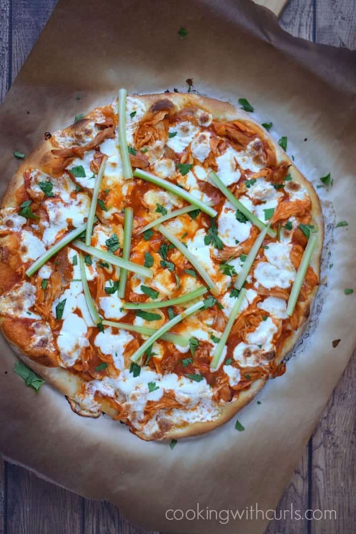 Looking down on a Buffalo Chicken Pizza topped with celery sticks on charred parchment paper.