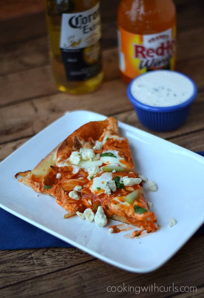 A slice of Buffalo Chicken Pizza topped with crumbled blue cheese and celery sticks, and a small bowl of ranch dressing in the background.