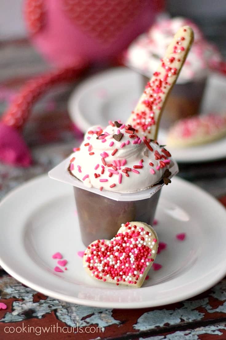 A chocolate pudding cup topped with whipped cream, red and pink sprinkles and a pie crust spoon on the side