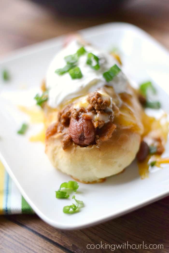 close up image of a chili and melted cheese covered hot dog with sour cream and sliced green onions on top sitting on a white plate