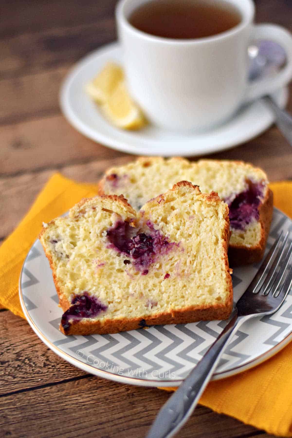 Two slices of lemon blackberry bread on a small plate with a cup of tea in the background.