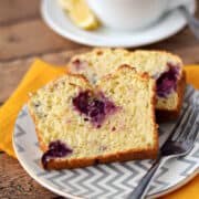 Two slices of lemon blackberry bread on a small plate with a cup of tea in the background.