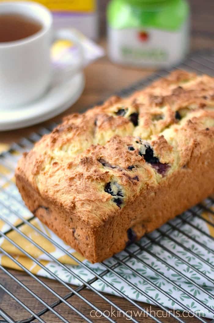 a loaf of lemon blackberry bread sitting on a wire rack with a cup of tea in the upper right corner