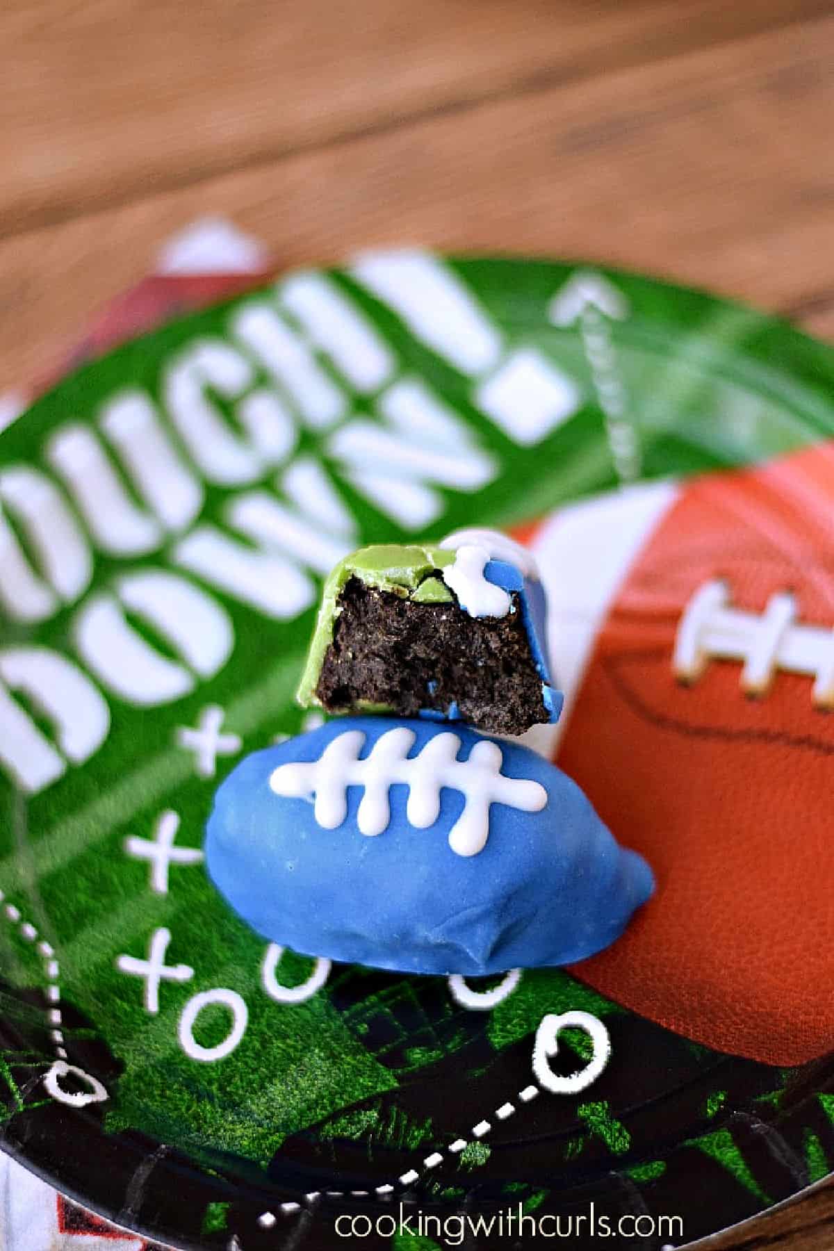 One blue football shaped cookie ball on a game day paper plate with a second cookie football bitten in half leaning against it. 