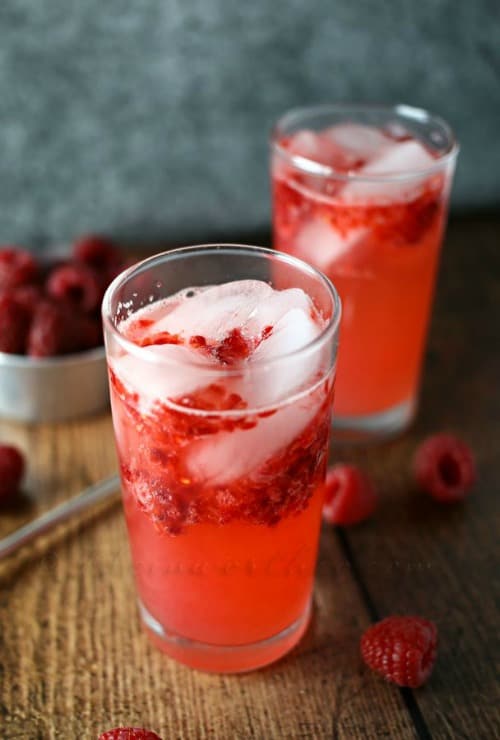 Two Sour Raspberry Tom Collins in highball glasses with raspberries scattered around the base.