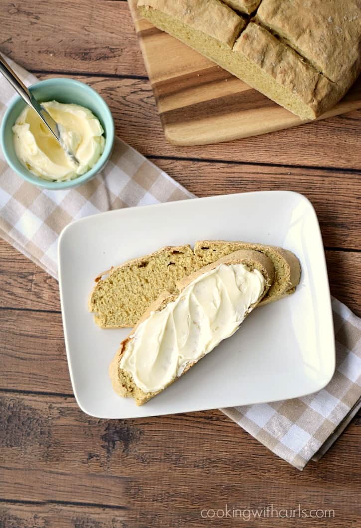 two slices of irish soda bread on a square, white plate covered in butter with a blue bowl of butter with a knife in the upper left corner and the remaining loaf of bread in the upper right on a wooden cutting board
