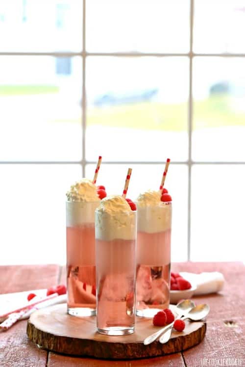 Three adult raspberry Italian cream sodas in tall, skinny glasses sitting on a wood circle in front of a window.