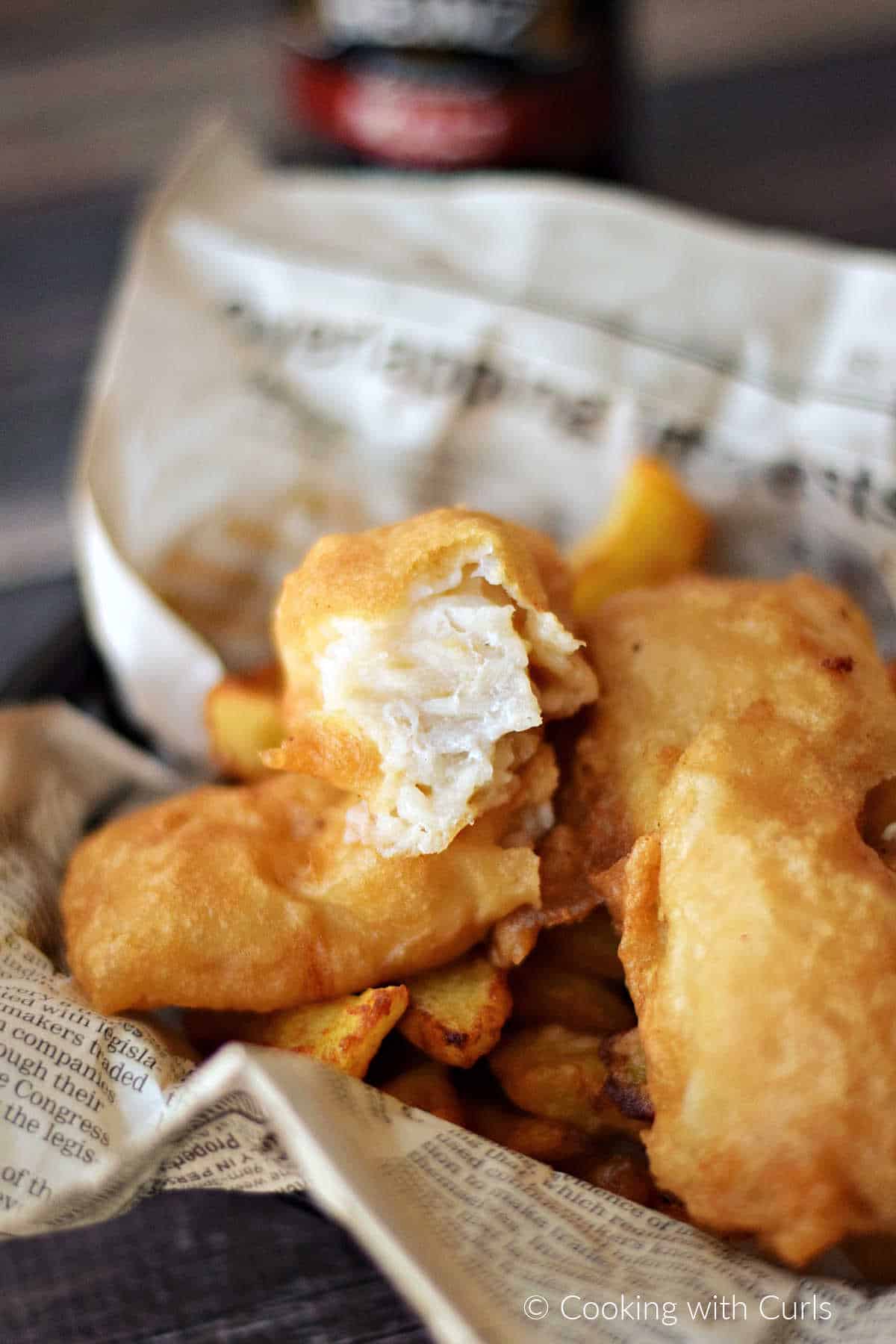 A broken piece of beer batter fish resting on a pile of fish and French fries.