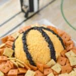 bacon cheddar cheeseball covered in shredded cheddar cheese and minced olives designed to look like a basketball sitting on a white plate surrounded by crackers