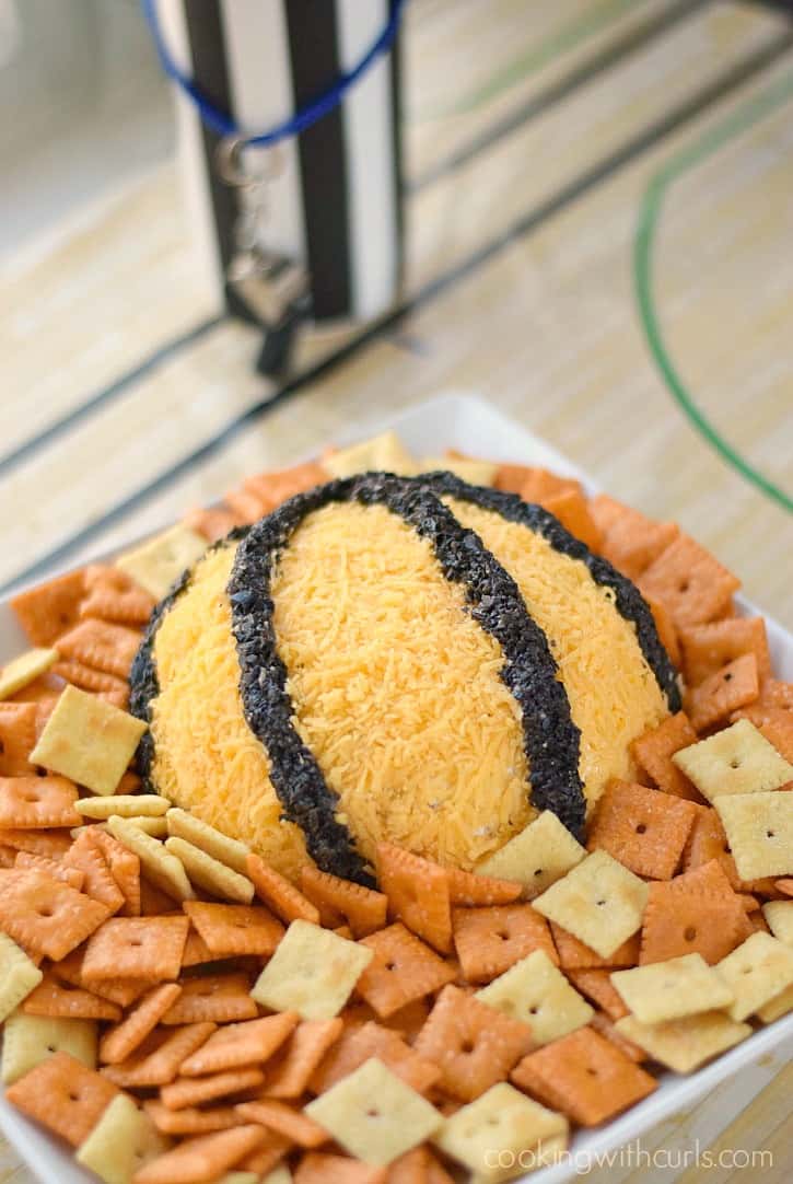 bacon cheddar cheeseball covered in shredded cheddar cheese and minced olives designed to look like a basketball sitting on a white plate surrounded by crackers