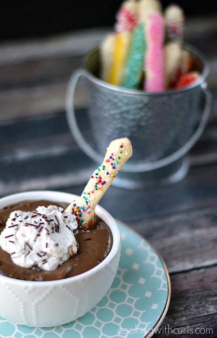 rainbow colored sprinkle covered pie crust spoon sticking out of a bowl of chocolate pudding with whipped cream and additional spoons in a metal pail up in the right hand corner