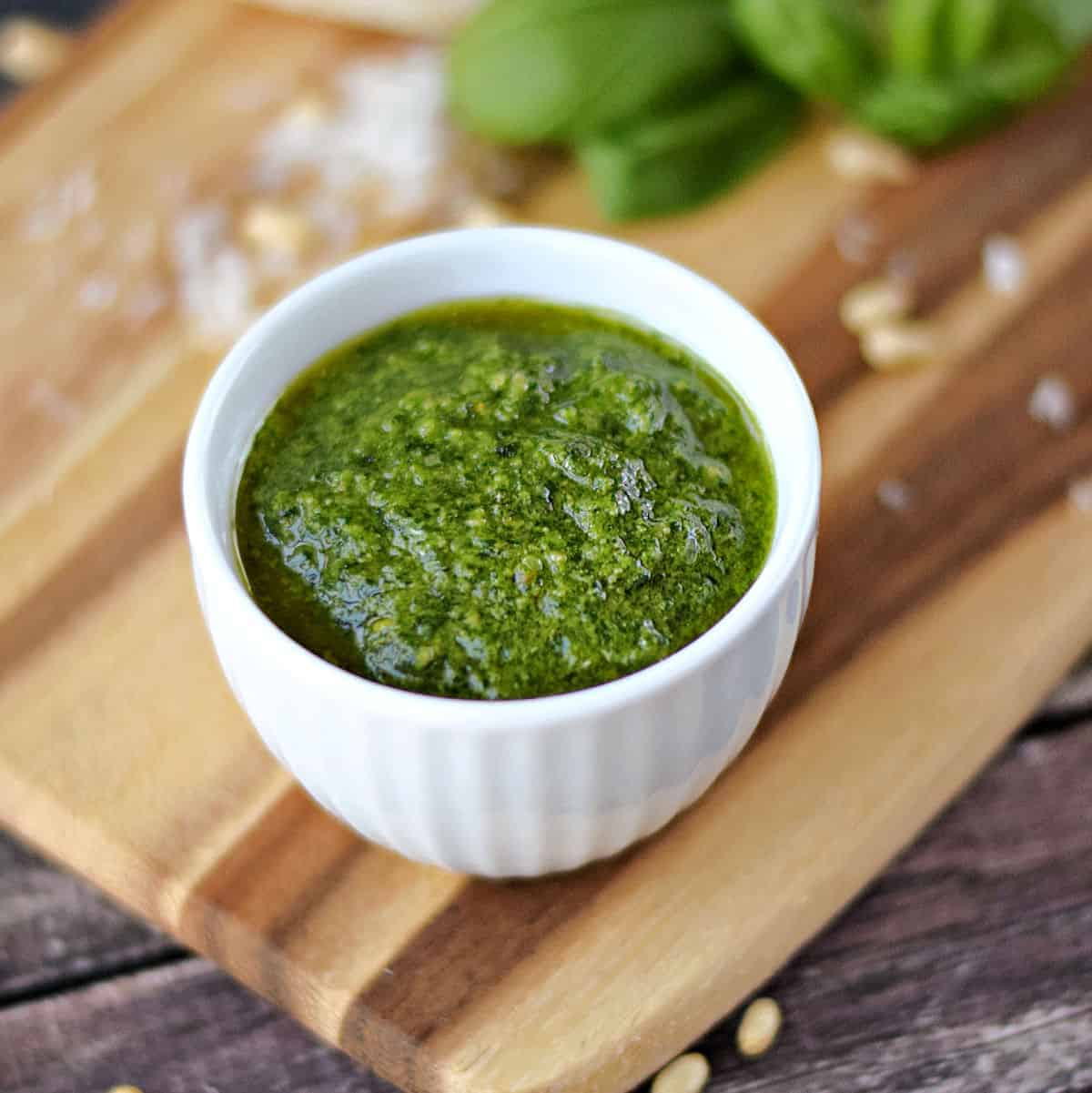 Basil Pesto in a small white bowl sitting on a wood cutting board that has been scattered with pine nuts, grated Parmesan and basil leaves.