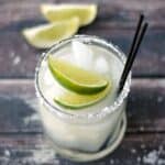 classic margarita in a small glass with two lime wedges and two black straws on a wooden board with two lime wedges in the background