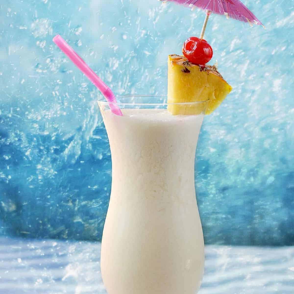A creamy white cocktail in a hurricane glass with a pink straw, pineapple wedge, cherry and pink paper umbrella.