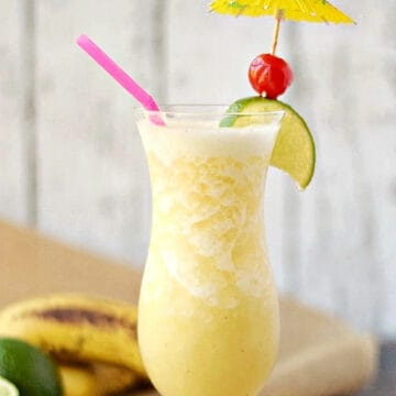 Frozen Banana Daiquiri in a hurricane glass garnished with a paper umbrella, lime wedge, and cherry.