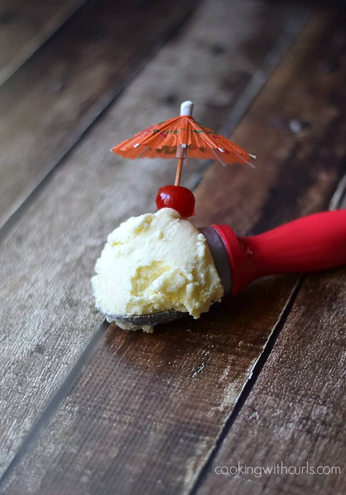 a scoop of Piña Colada Ice Cream in a pink ice cream scoop with a cherry and orange paper umbrella sticking out of the side.