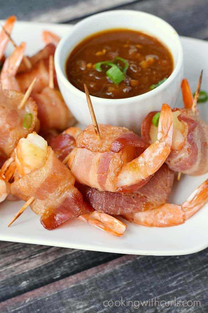 A stack of bacon wrapped shrimp and pineapple on a plate with a bowl of teriyaki sauce.