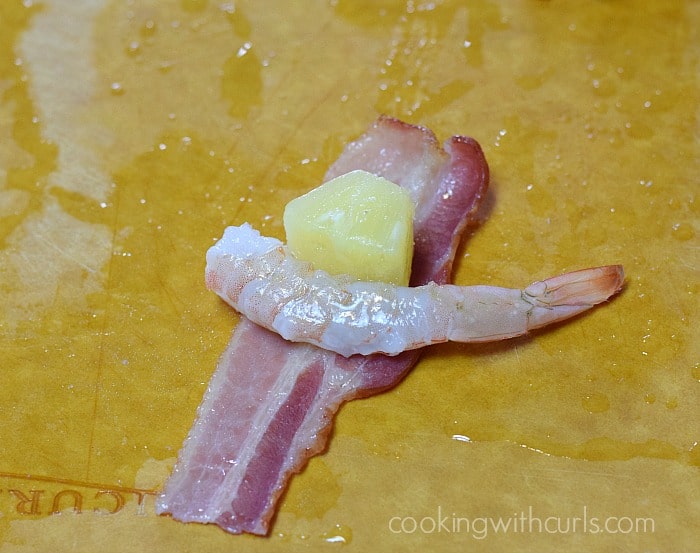 A piece of shrimp and a pineapple chunk laying on half a piece of bacon on a cutting board.