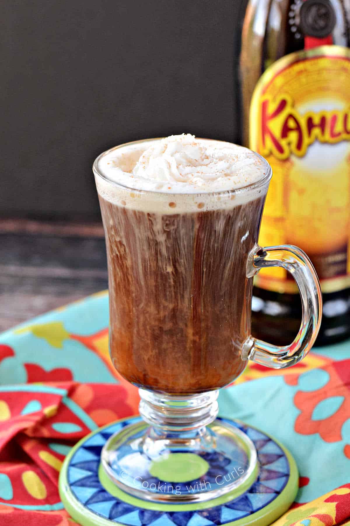 A coffee cocktail in a tall glass mug topped with whipped cream, with a bottle of Kahlua in the background.