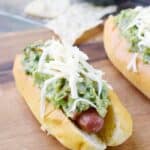 two hot dog buns sitting on a wooden board filled with a hot dog and guacamole, topped with shredded cheese with tortilla chips in the back ground
