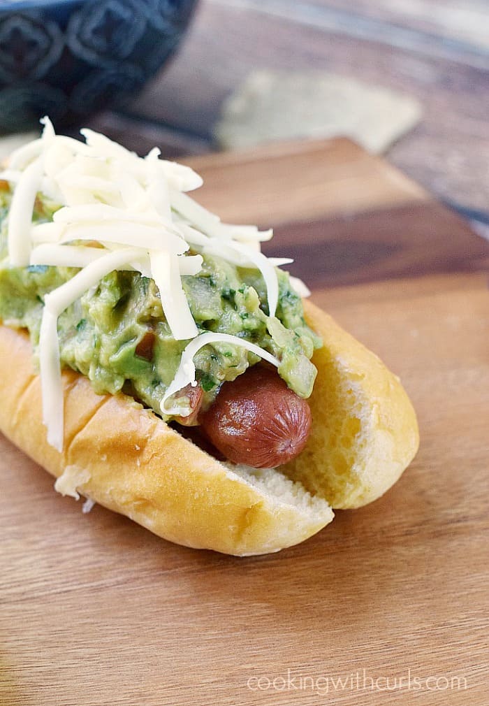 guacamole and shredded cheese topped hot dog in a bun sitting on a wooden board with tortilla chips in the background