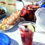 a clear plastic cup filled with berries and sangria surrounded by cilantro lime shrimp, a pitcher of sangria, blue plates and bottles of wine