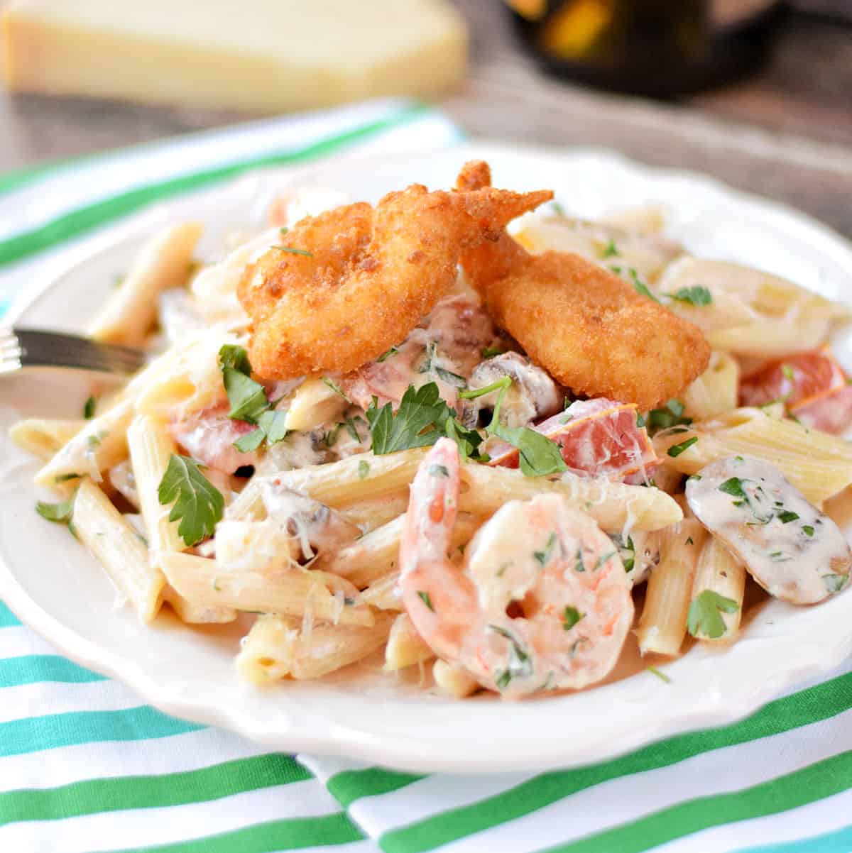 Penne pasta mixed with a creamy shrimp sauce, shrimp, mushrooms, tomatoes, and topped with three breaded shrimp on a white plate.