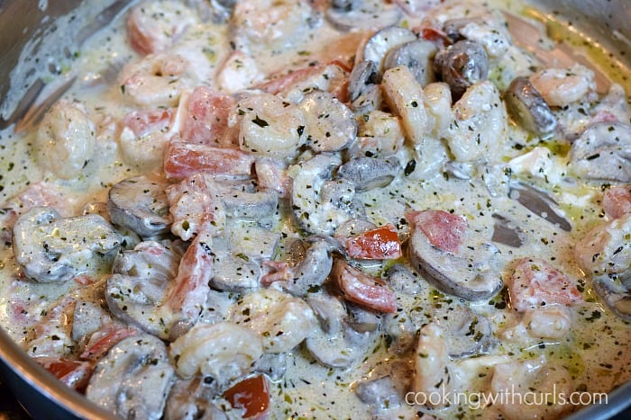 Creamy sauce mixed with shrimp, sliced mushrooms, and chopped tomatoes in a skillet.