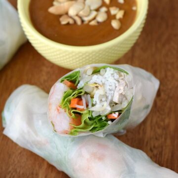 spring rolls laying on a wooden board with a diagonal cut half laying on top and a yellow bowl of peanut sauce in the left corner