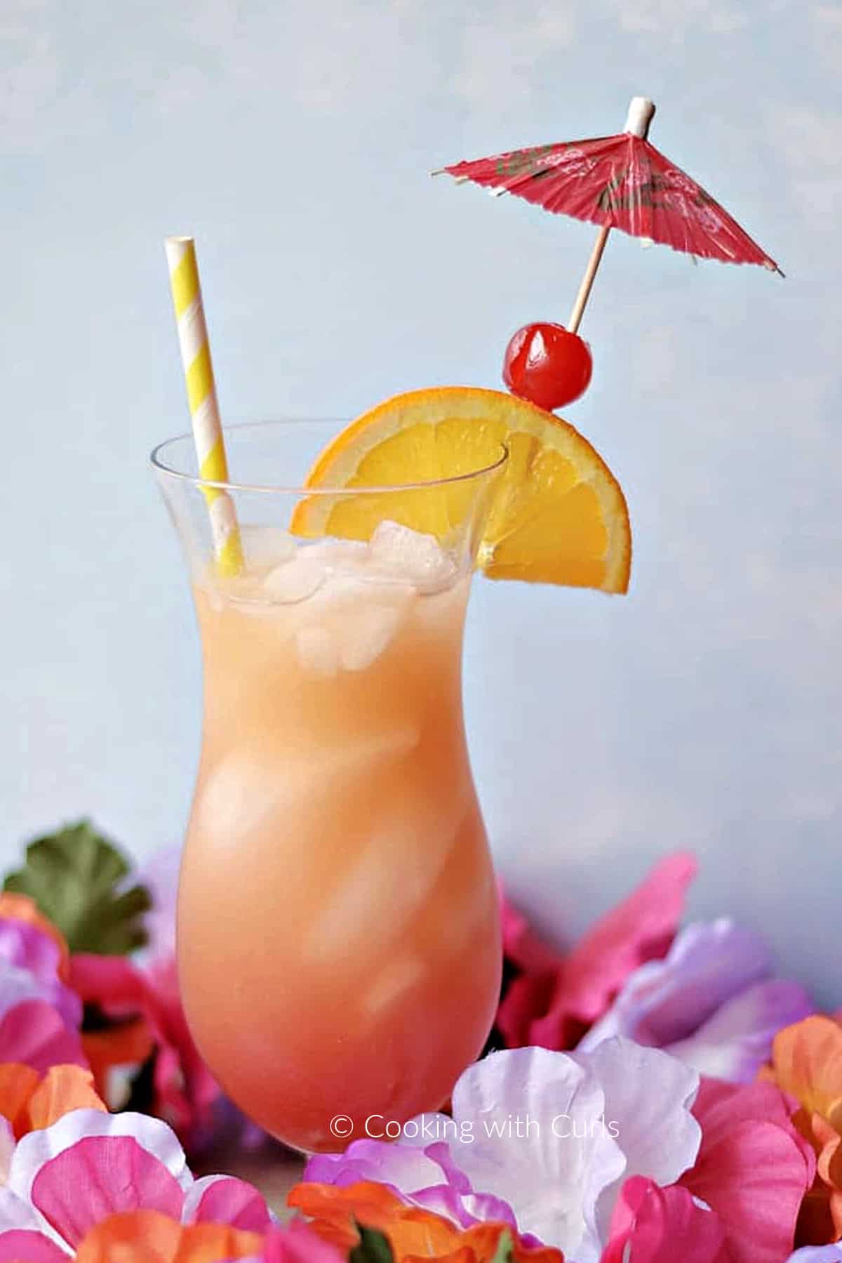 Bahama Mama drink garnished with an orange slice, cherry, and paper umbrella surrounded by colorful flowers. 