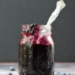 glass jar filled with blueberry preserves with a spoon sticking out of the side and surrounded by fresh blueberries
