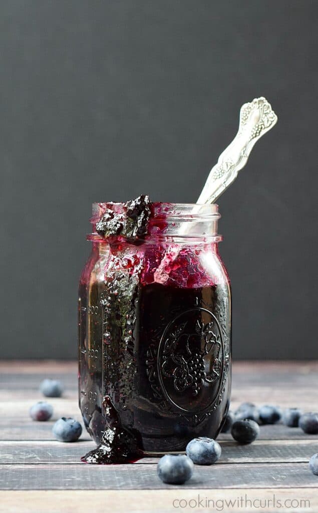 glass jar filled with blueberry preserves with a spoon sticking out of the side and surrounded by fresh blueberries
