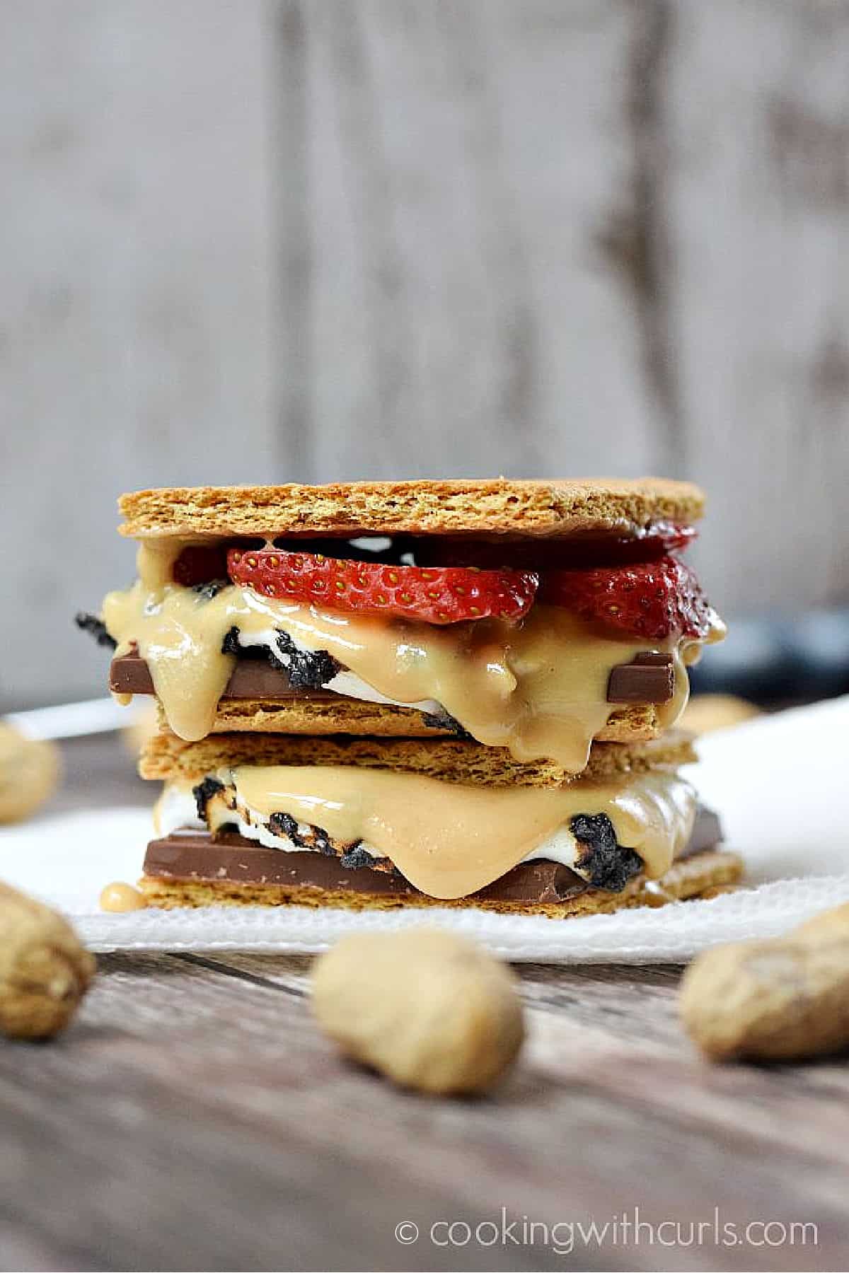 A chocolate bar, toasted marshmallows, peanut butter, and sliced strawberries between graham crackers stacked on a napkin. 