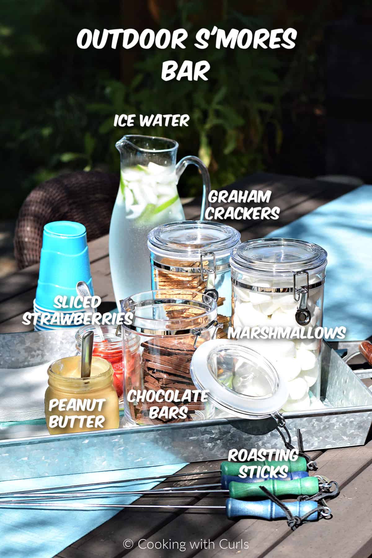Glass canisters with marshmallows, graham crackers, chocolate bars, peanut butter, and sliced strawberries on a metal tray with a pitcher of water in the background. 