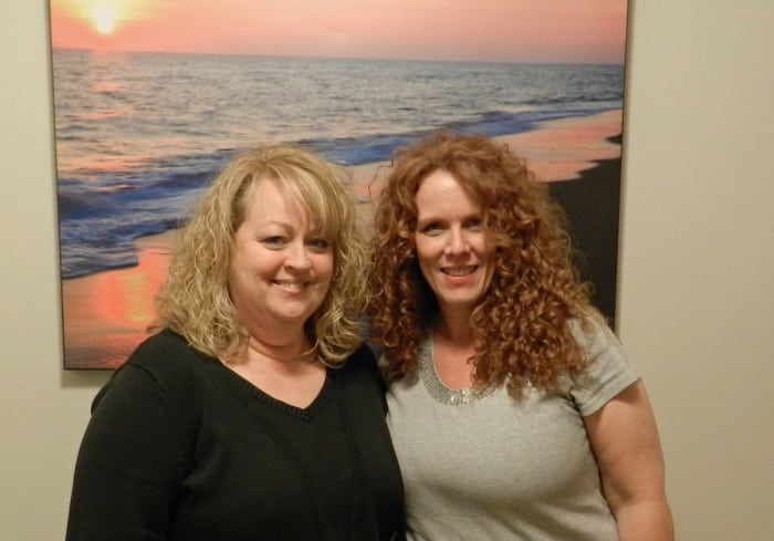 One woman with curly blonde hair and a second with curly read hair standing in front of an ocean painting.