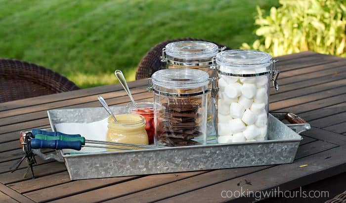 Glass canisters with marshmallows, graham crackers, chocolate bars, peanut butter, and sliced strawberries, and toasting sticks on a metal tray. 