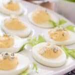 buffalo style eggs topped with blue cheese crumbles and surrounded by celery leaves on a white egg platter
