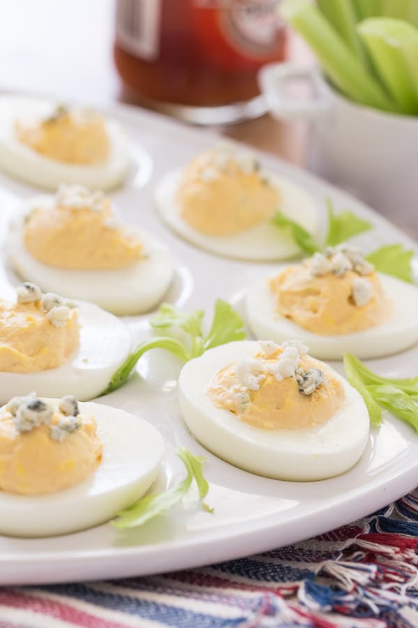 buffalo style eggs topped with blue cheese crumbles and surrounded by celery leaves on a white egg platter 