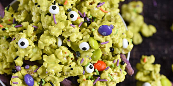 A bowl overflowing with green candy coated popcorn, sprinkles, purple pretzel sticks, and multi-colored candy coated chocolates.