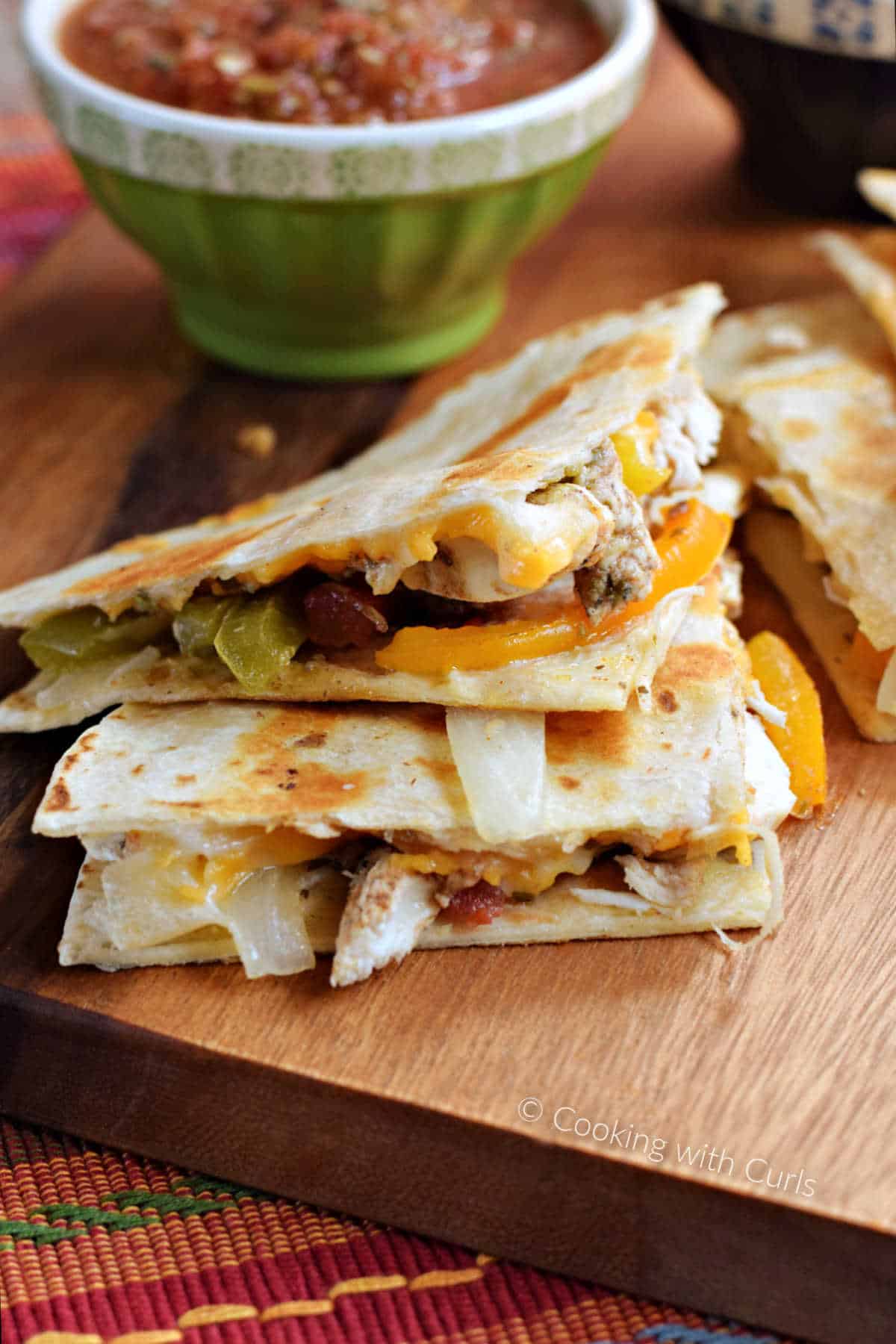 Two chicken fajita quesadilla wedges stacked on a board with a bowl of salsa in the background.