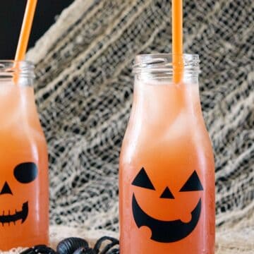 beige netting draped over a black background with two glass bottles decorated with jack o lantern faces, filled with orange cocktails and orange straws