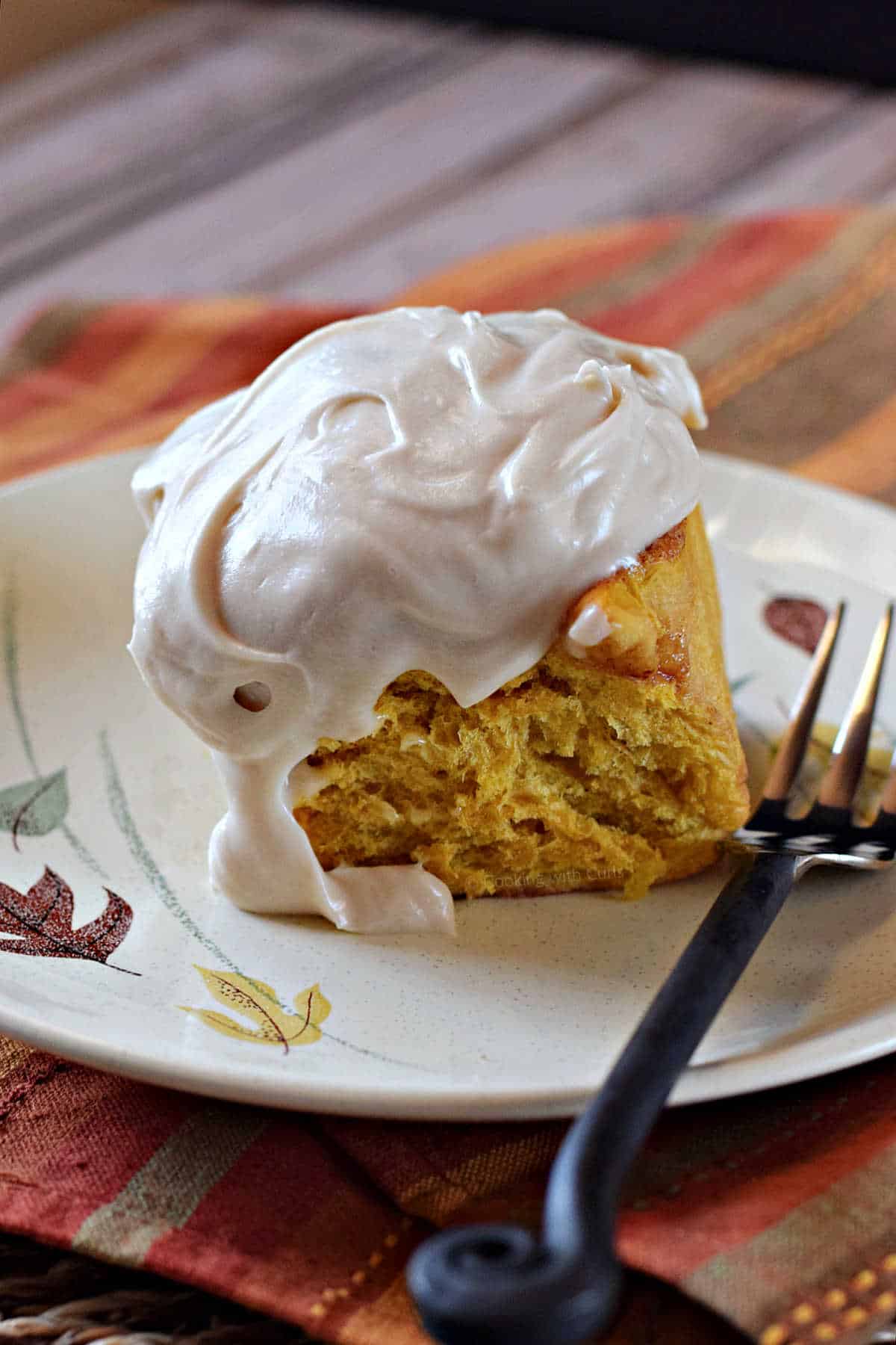 A pumpkin cinnamon roll topped with cream cheese frosting on a plate with a fork on the side.