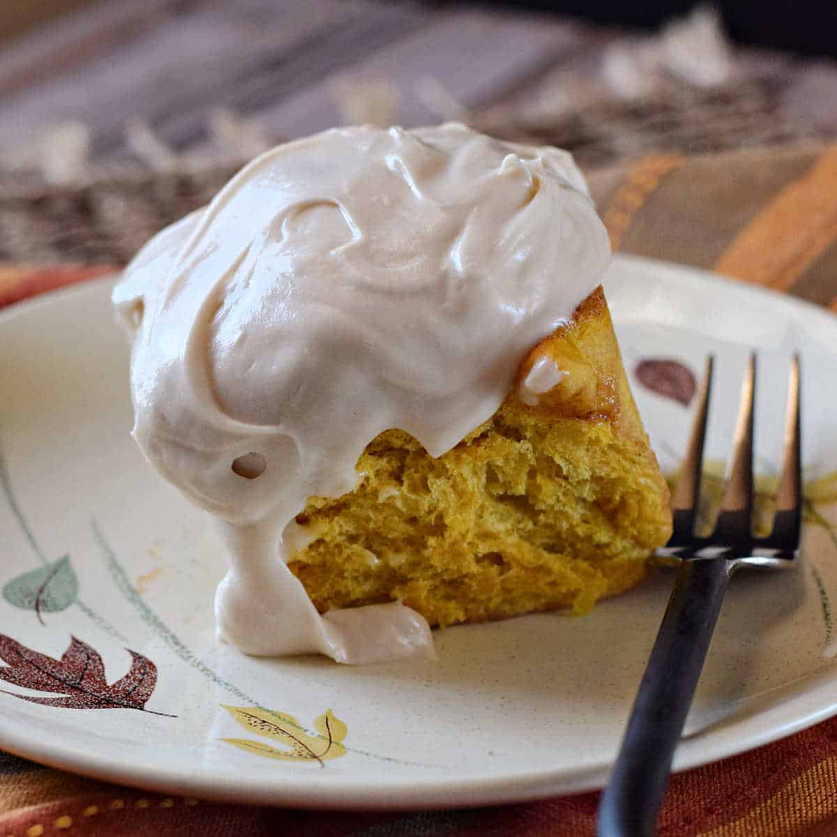 A pumpkin cinnamon roll topped with cream cheese frosting on a plate with a fork on the side.