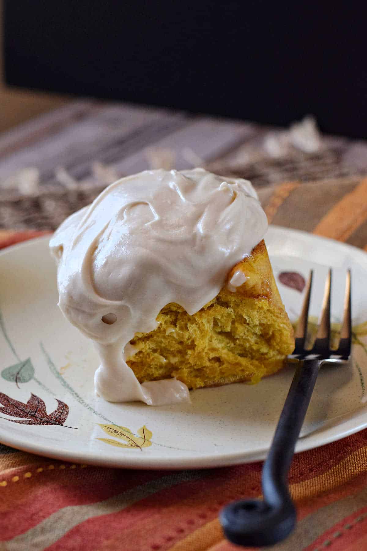 A pumpkin cinnamon roll topped with cream cheese icing on a plate with a fork on the side.