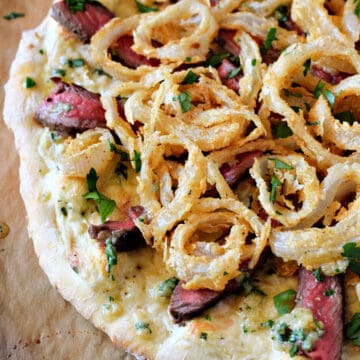 Pizza topped with white pizza sauce, strips of medium-rare steak, Gorgonzola butter, and crispy onion rings.