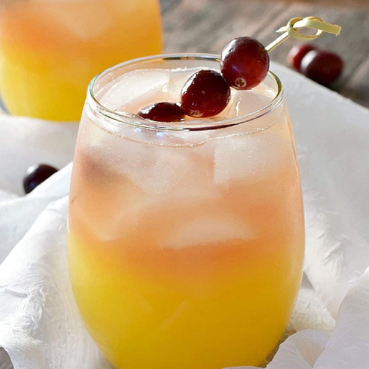 two small, clear glasses with orange on the bottom and peach colored on the top filled with ice cubes and garnished with cranberries on a bamboo stick.