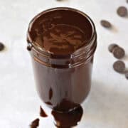 Hot fudge sauce overflowing a glass jar surrounded by chocolate chips.